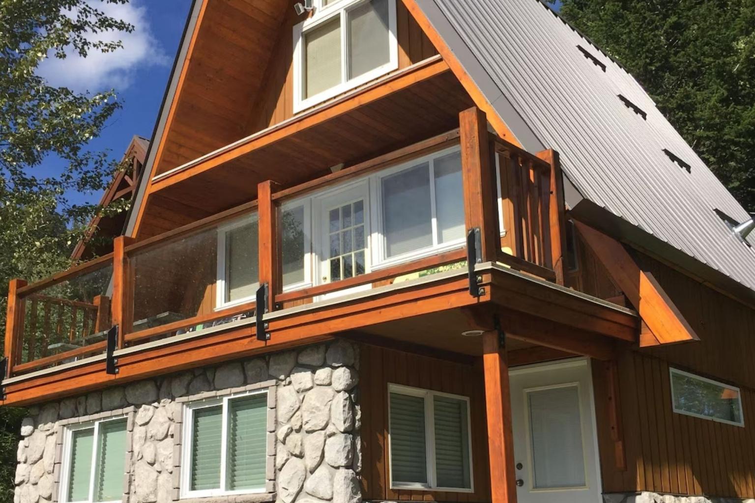 A-frame cabin with wood balcony and stone lower walls among trees