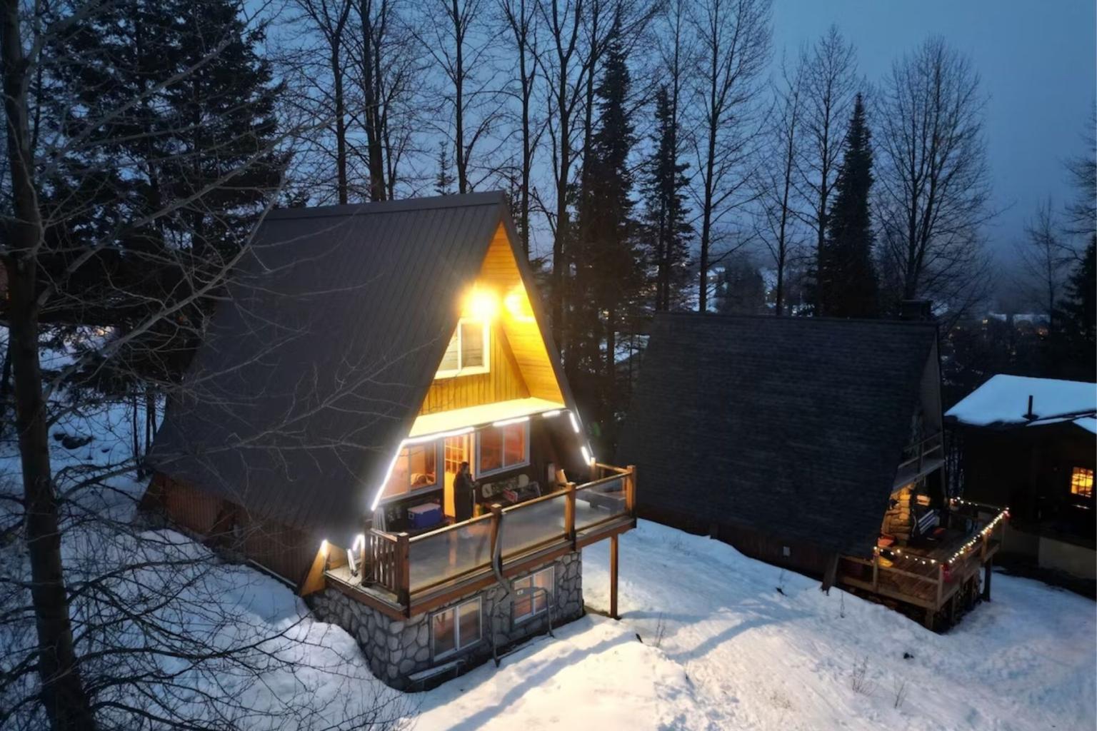 Snow-covered A-frame cabin glowing warmly at dusk among tall trees.