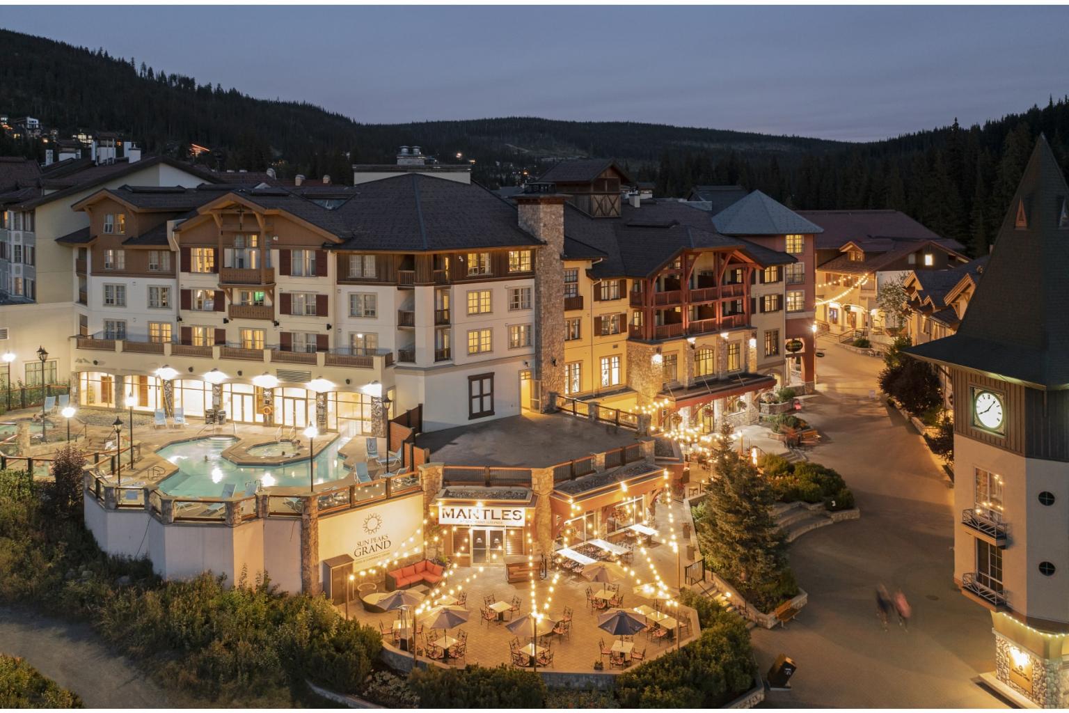 Twilight view of a warmly lit alpine hotel village with outdoor dining terraces.