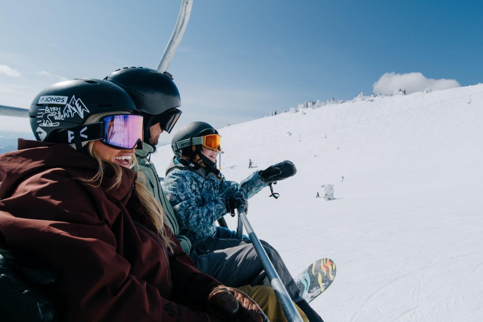 Two snowboarders in helmets and goggles riding a chairlift over snowy mountains.