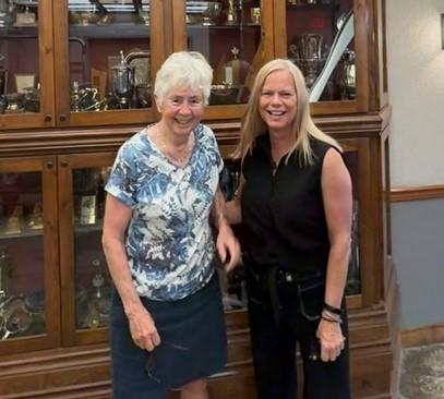 Two smiling women standing in front of a glass display cabinet.