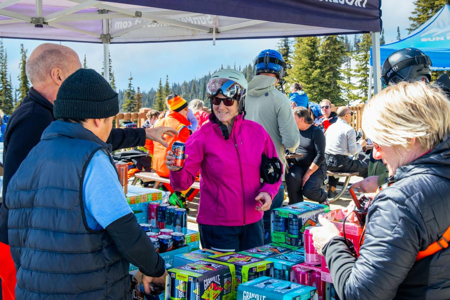 A skier being handed a cold can of beer at a table full of beer cases in an outdoor setting surrounded by many other people. 