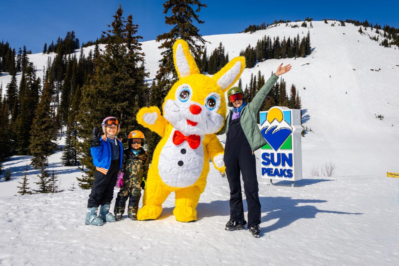 Skiers pose with a cute yellow bunny rabbit mascot on a snowy mountain.