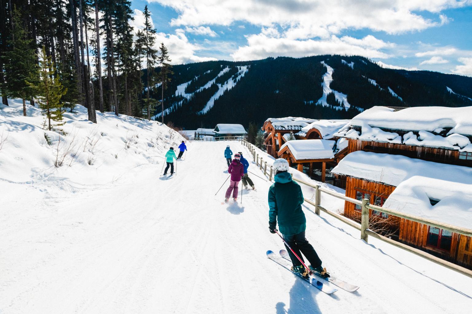 Skiers on a snowy trail beside a mountain lodge under blue skies.
