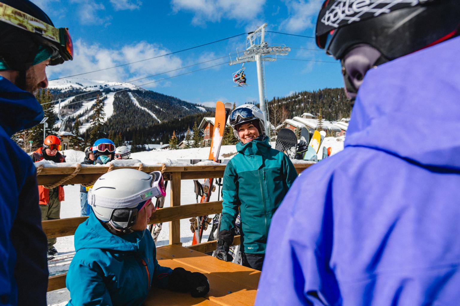 Skiers in colorful winter gear gather at a picnic table with Tod mountain and chairlift in background.