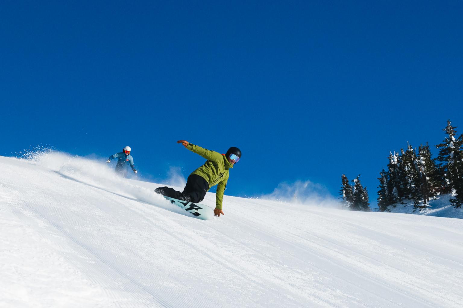 snowboarder carving down groomed slope with bright blue sky and skier trailing in background