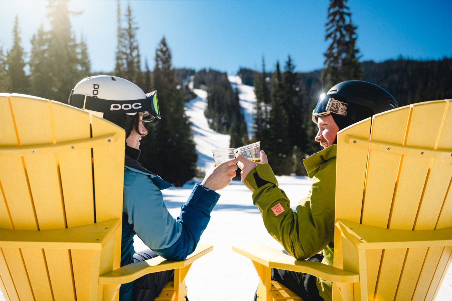 Two skiers toast with mugs between yellow chairs on a sunny snowy mountain.