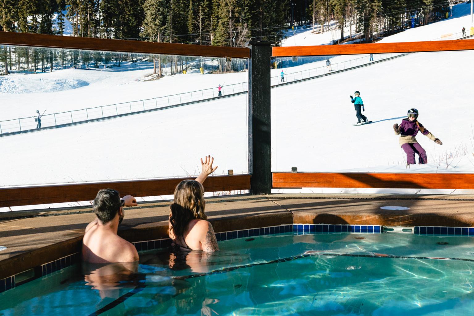 parents waving at their kids from a hot tub while kids ski and snowboard by and wave back