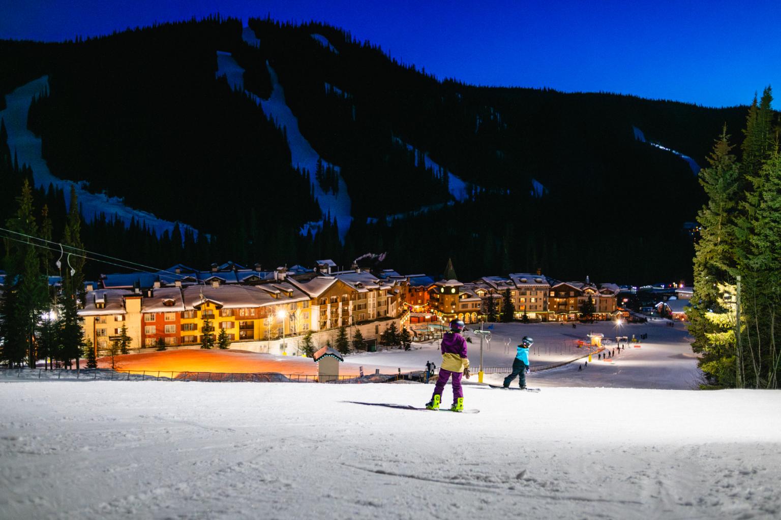 Two kids skiing together at night on lit slope above sun peaks village in winter