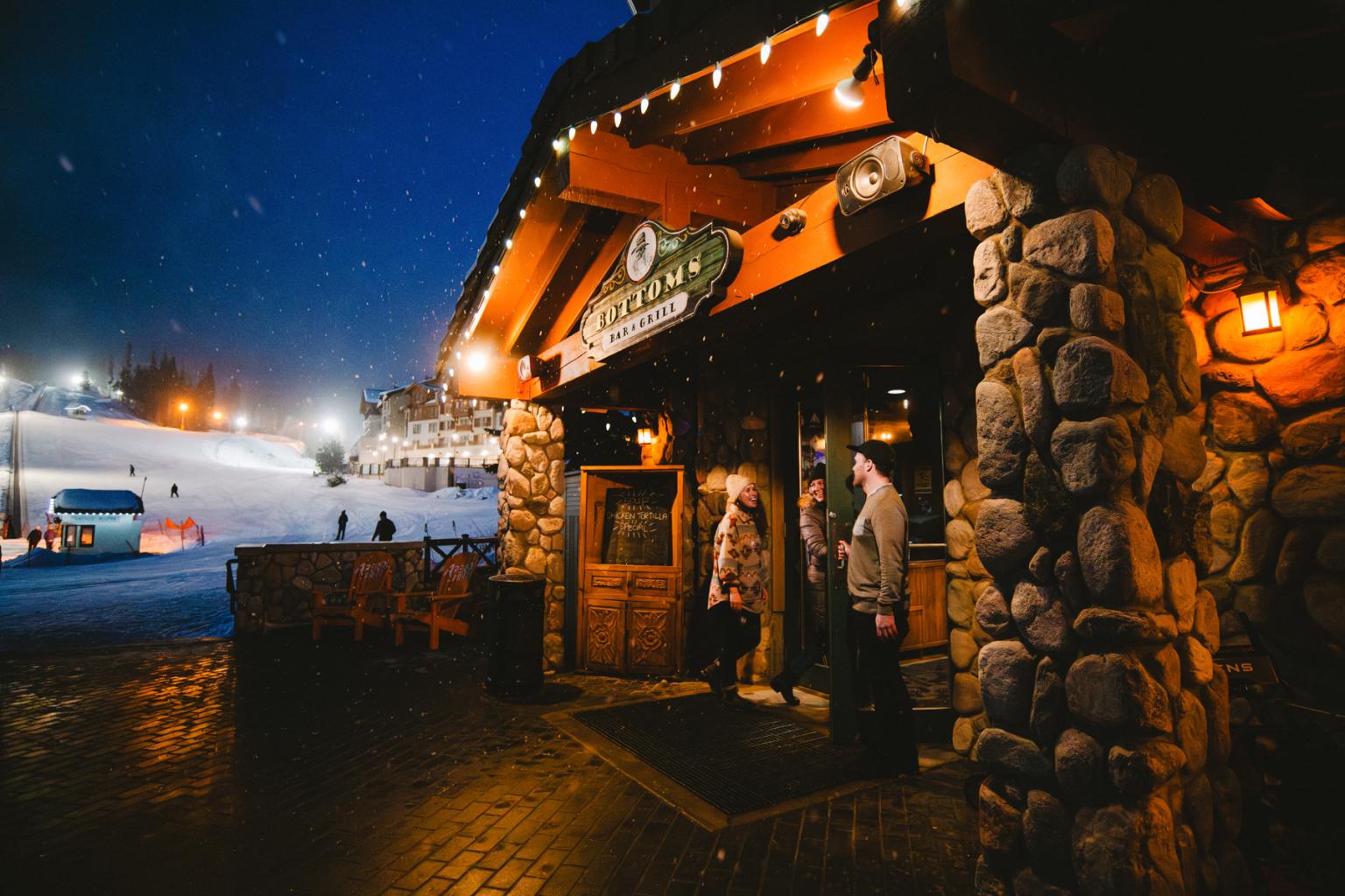 Man holds door open for two women entering Bottoms Bar at the bottom of a ski run in Sun Peaks at night with snow falling lightly