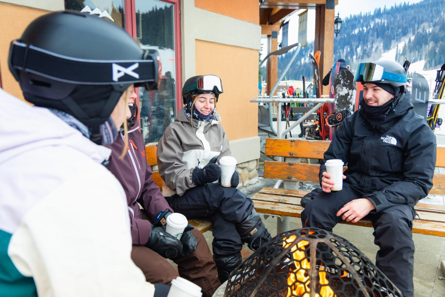 Skiers in helmets gather around a fire pit outside a mountain lodge.