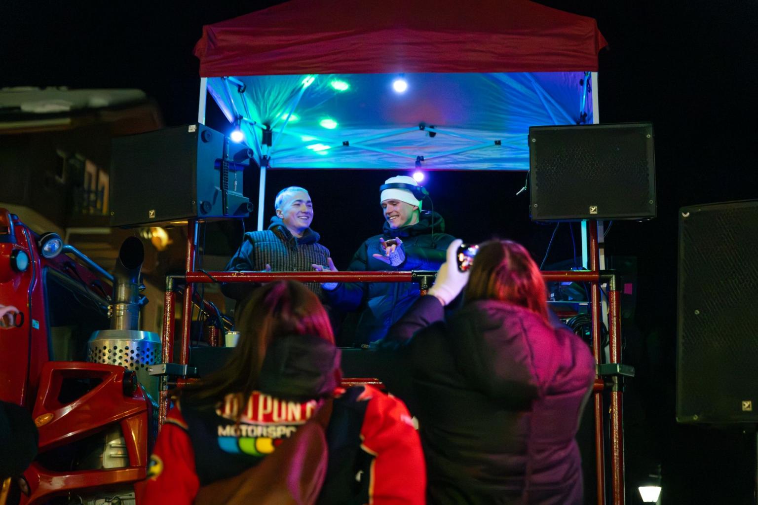 Nighttime outdoor music performance on a disco snowcat with two DJs lit with blue stage lights under a red tent.