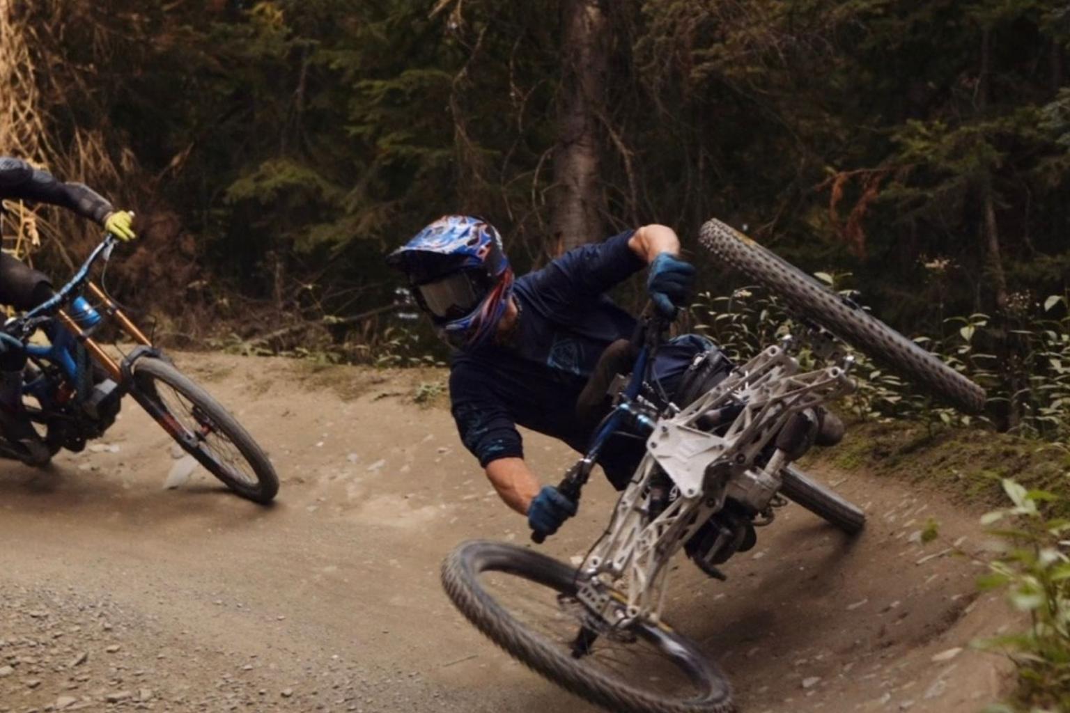 Mountain biker leaning into a dirt trail turn, with another rider behind.