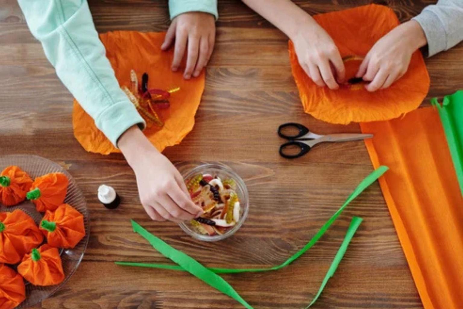 Children making orange paper pumpkins at a craft table with scissors and ribbon.