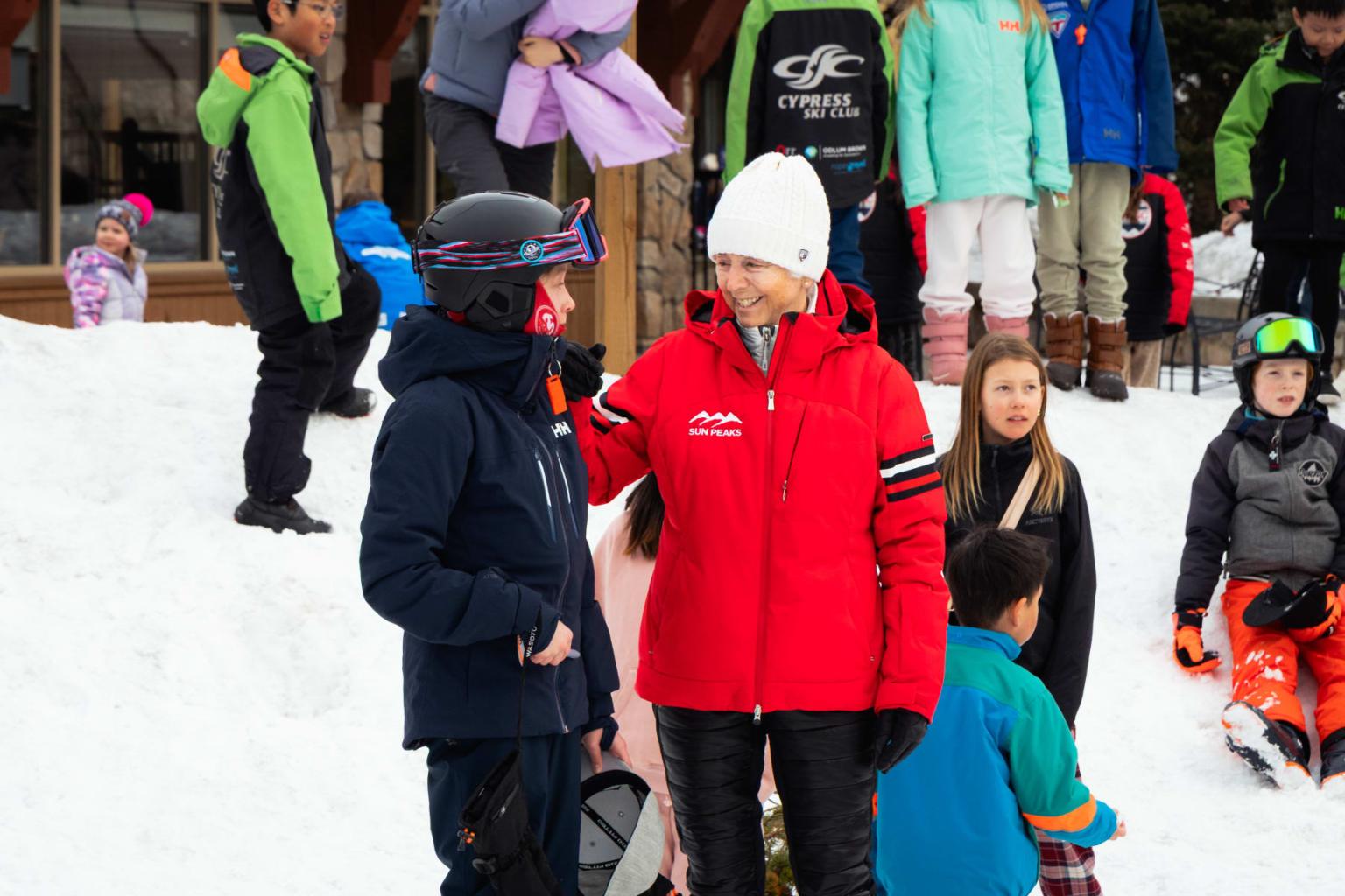 Woman in red ski jacket and white beanie (Nancy Greene) stands on a snowy slope with a child dressed in black ski gear..