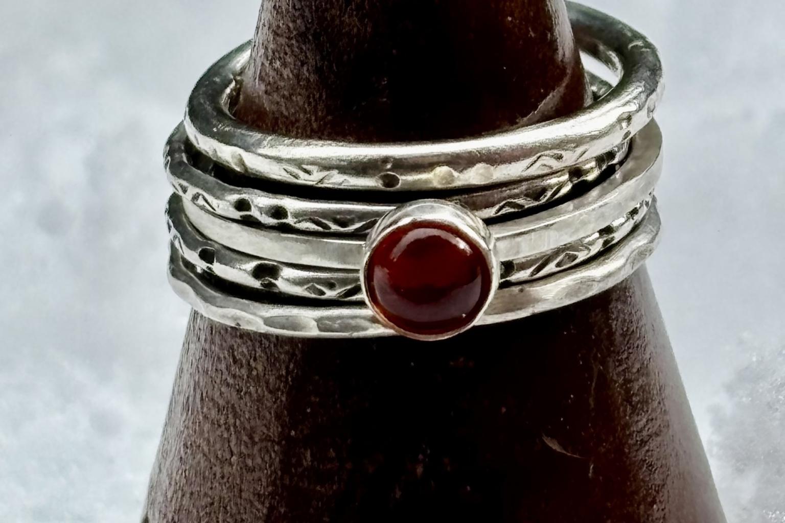 Stacked silver rings with textured bands and a red gemstone on a dark cone.