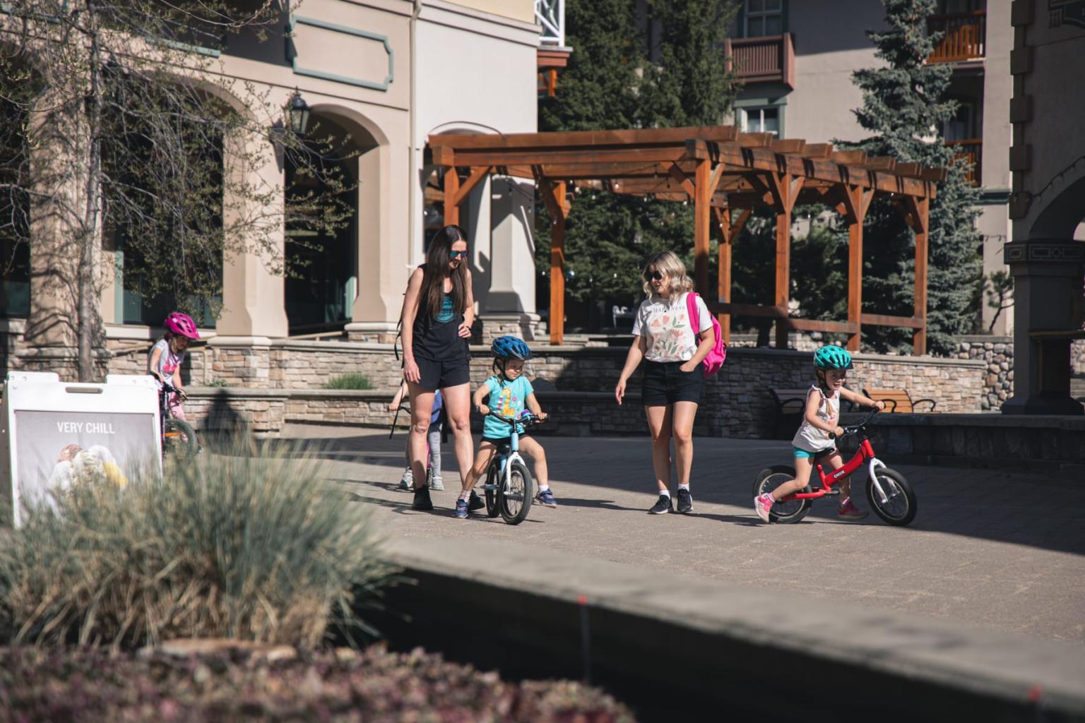 Woman and children walking past an outdoor café patio on a sunny street.