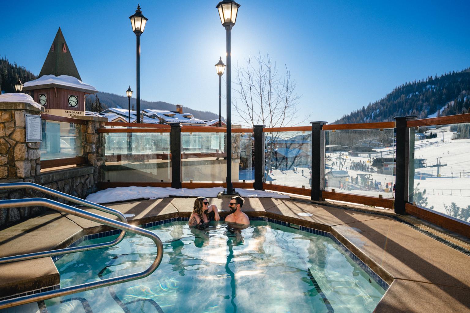 A couple sitting in a sunny sun peaks hot tun with clock tower and chairlifts in background