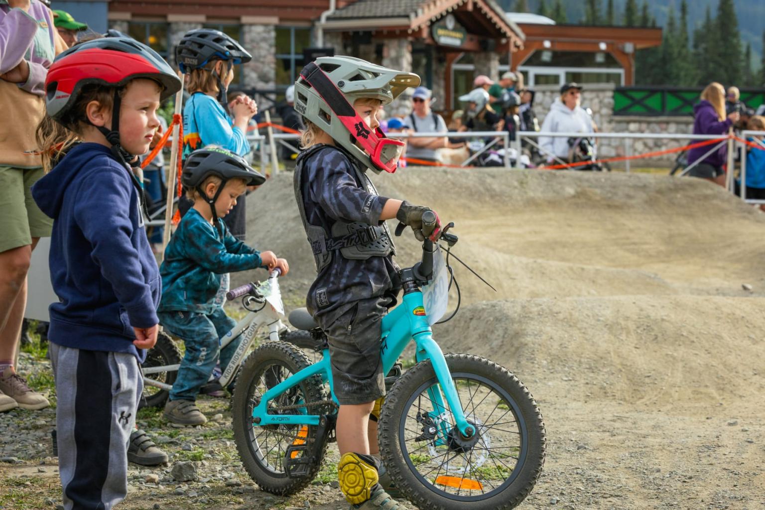 Child in helmet riding a turquoise bike at a muddy motocross track.