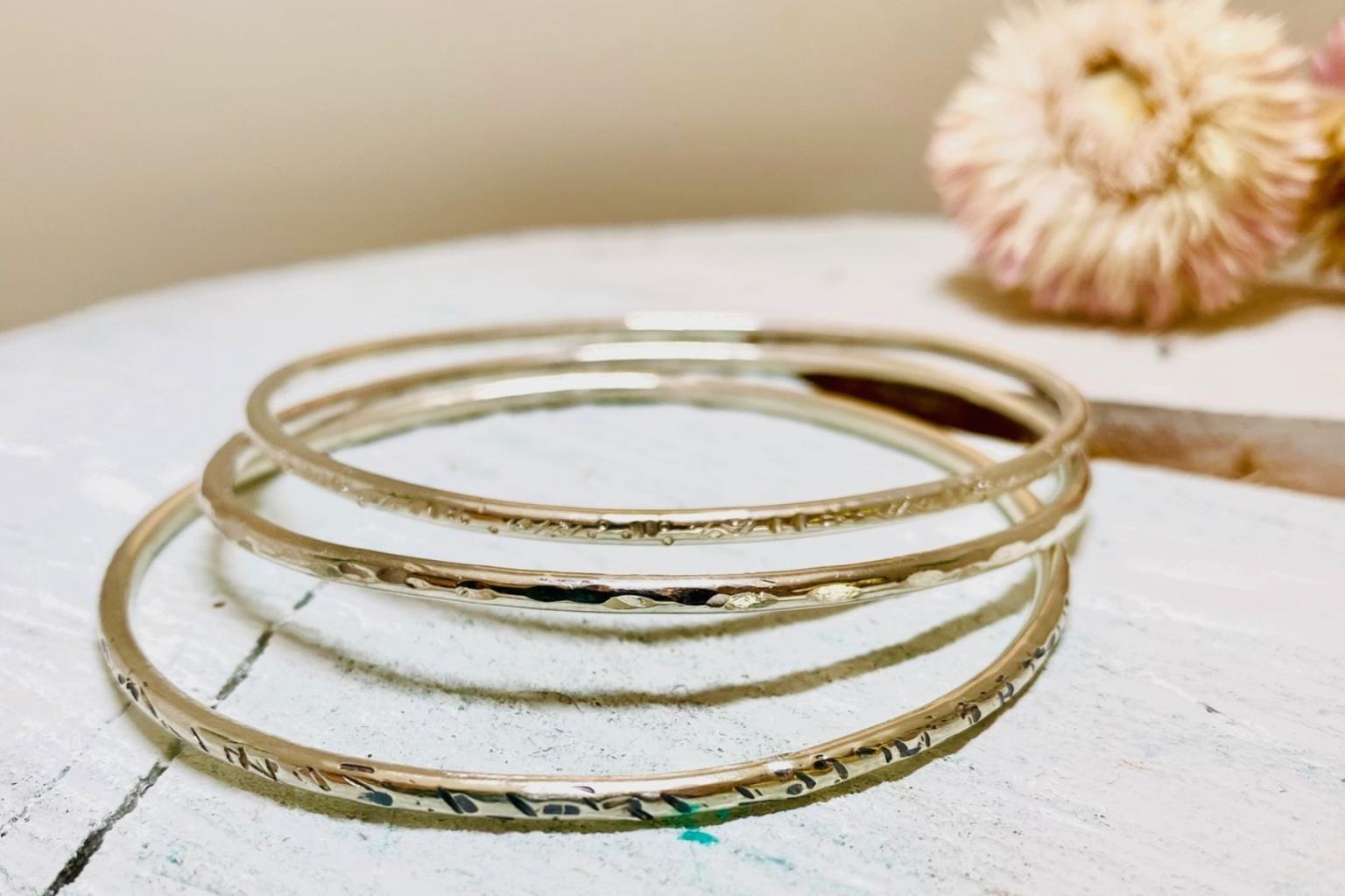 Three thin gold bangles on a white wooden surface beside a dried flower.