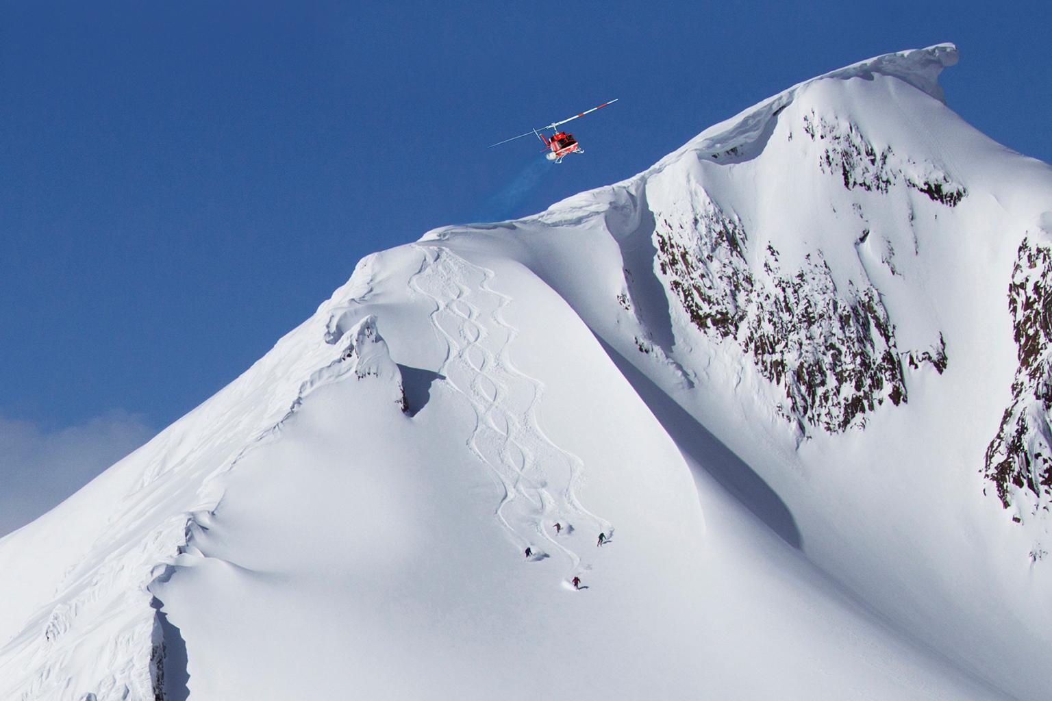 A helicopter flying above a mountain with skier riding down the powder