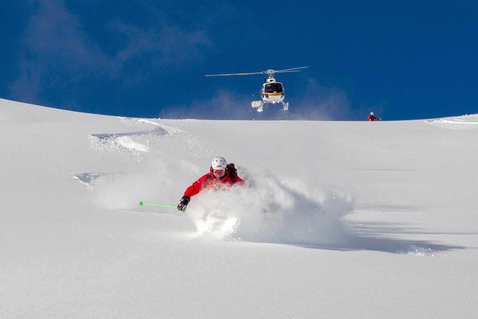 A skier riding down in powder with blue skies in the background and a helicopter following them