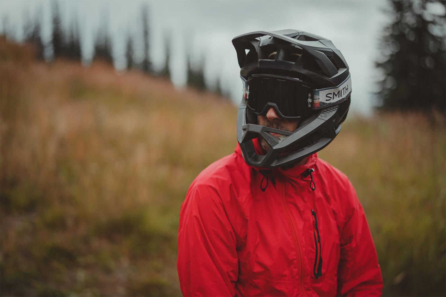 Mountain biker as seen from the chest up, looking slightly off camera with a red jacket and a black full face helmet and goggles