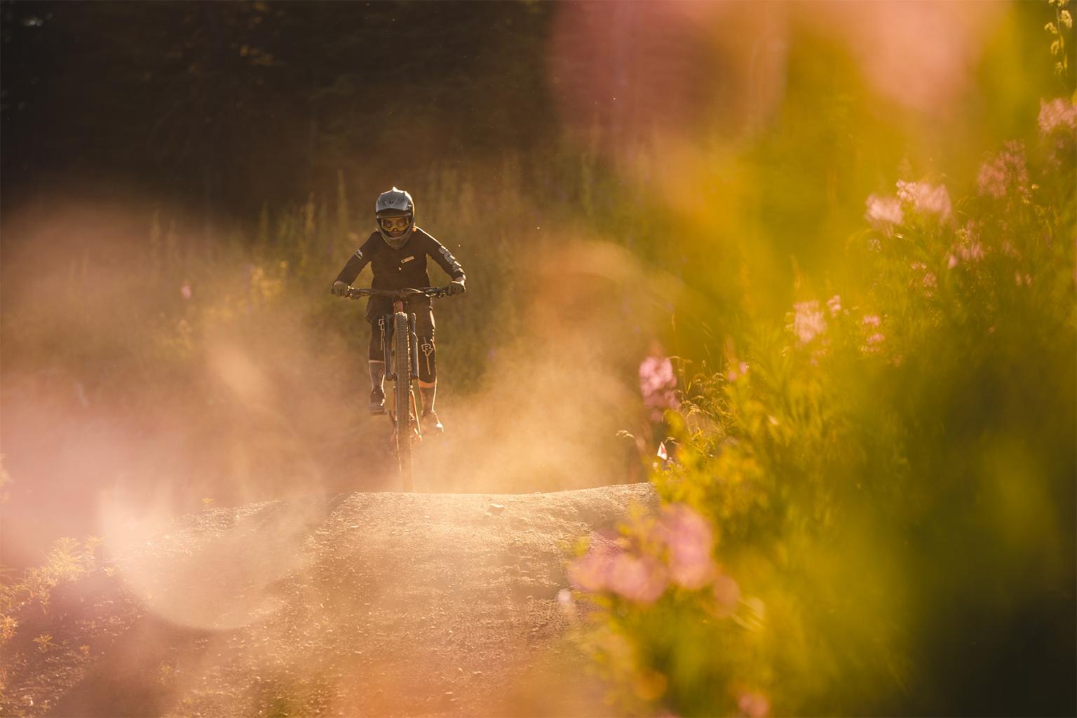 Mountain biker rides over a jump with tall grass and flowers to either side 
