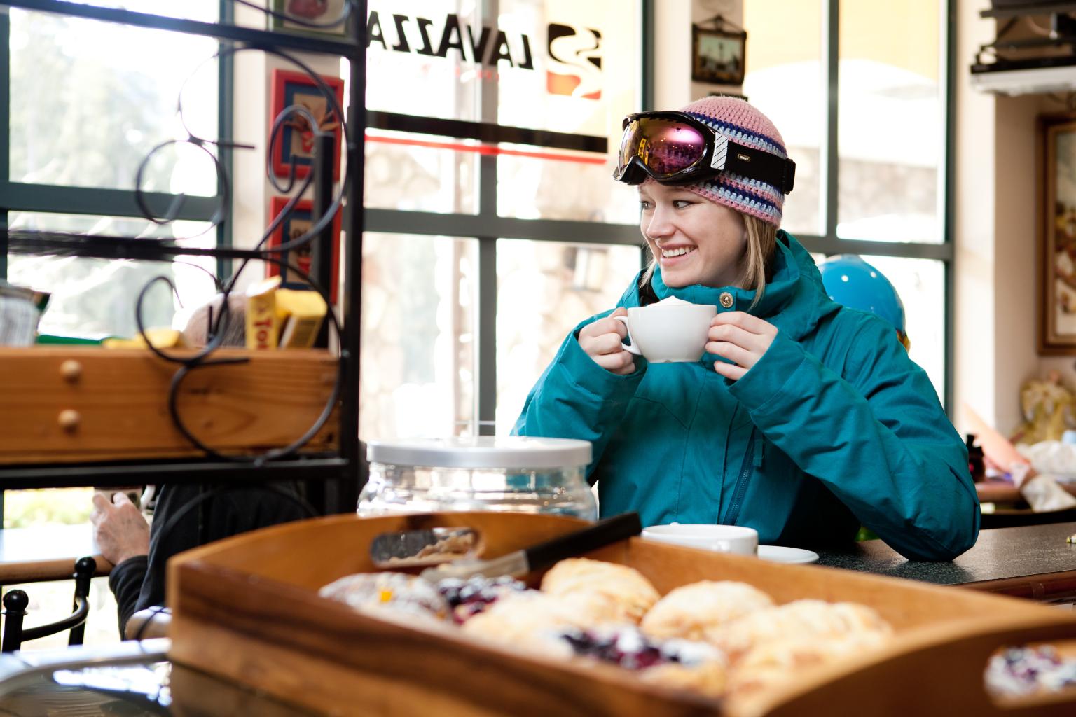 A woman dressed in ski gear drinking coffee from a mug while sitting inside a coffee shop