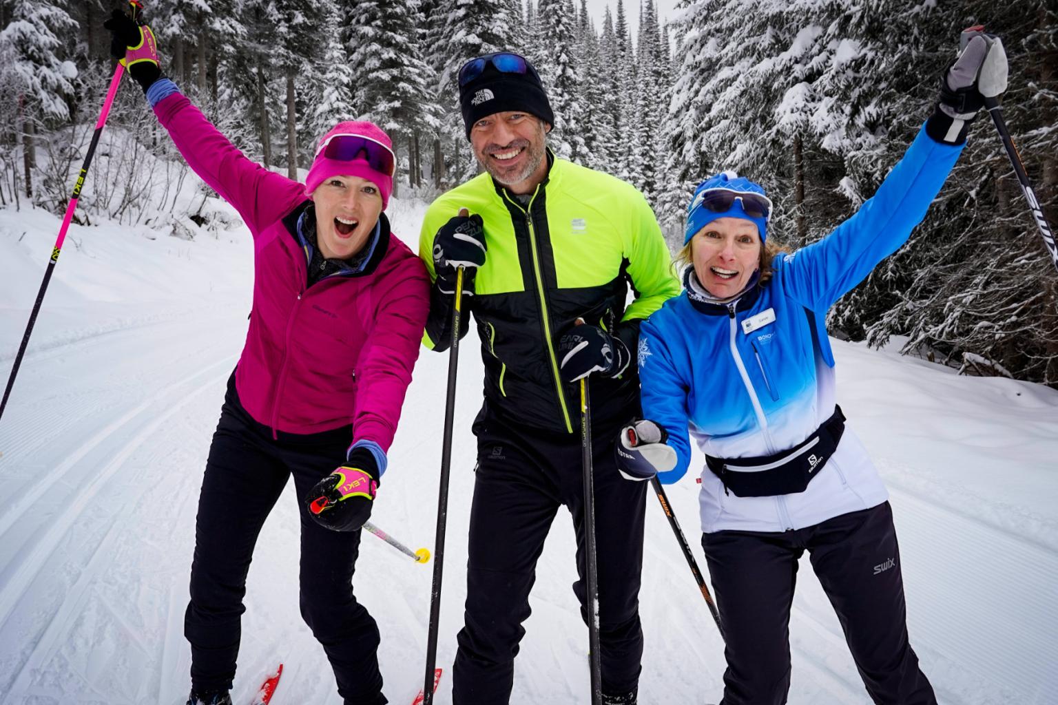Three Nordic skiers smiling to the camera with snow covered trees around them