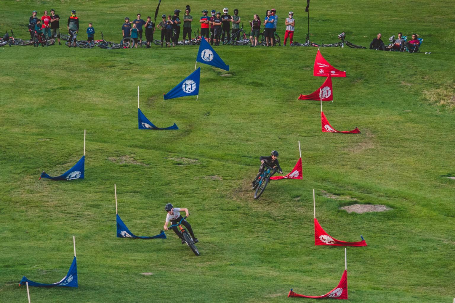 Two bikers on a grass dual slalom track with people sitting above them