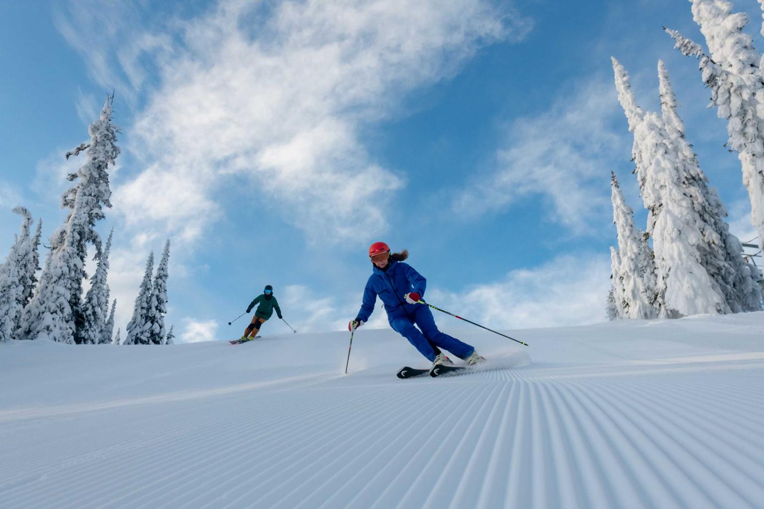 Two skiers riding down the hill on freshly groomed snow with blue skies and trees in the background