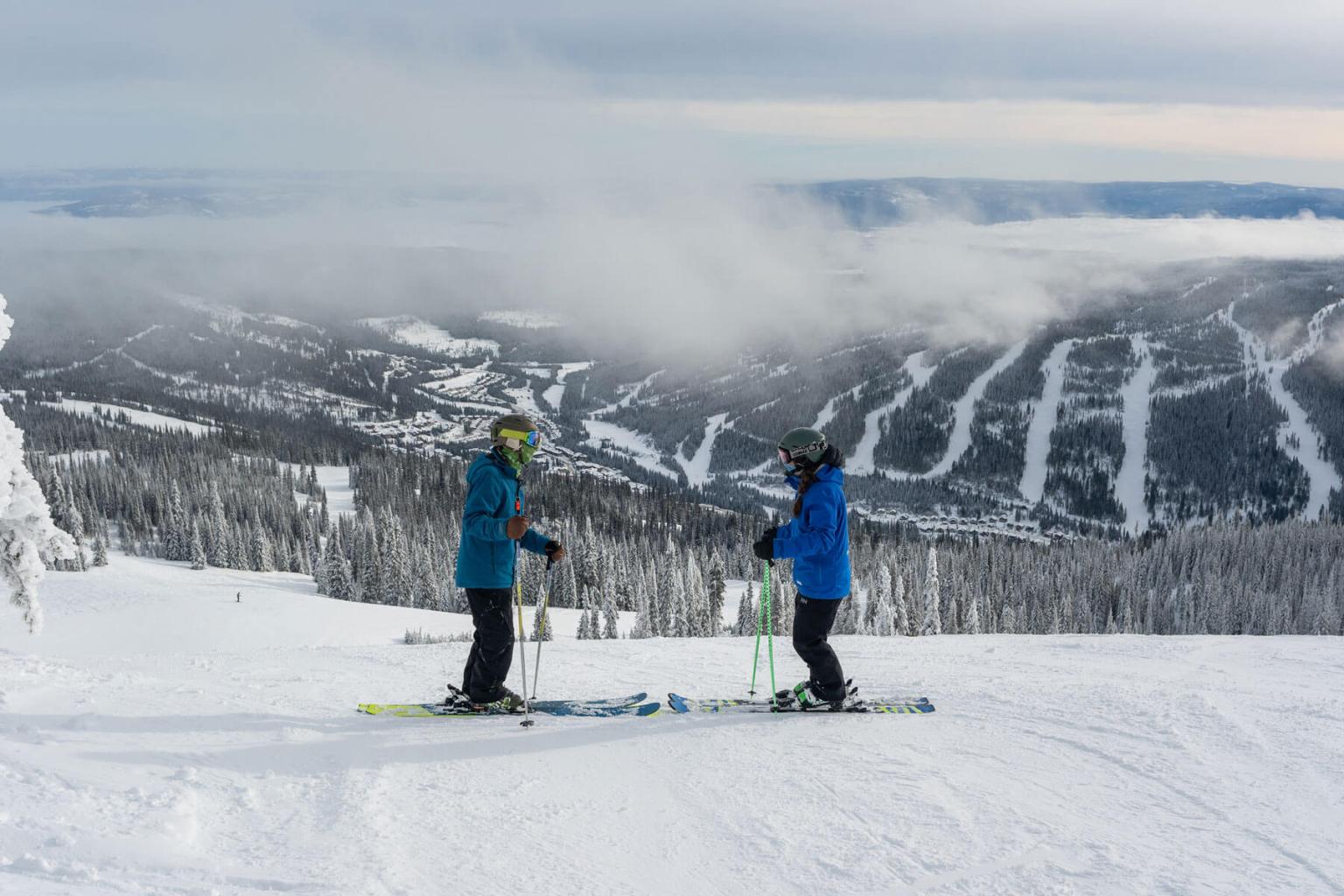 Two skiers facing eachother while standing at the top of a run with mountain views in the distance
