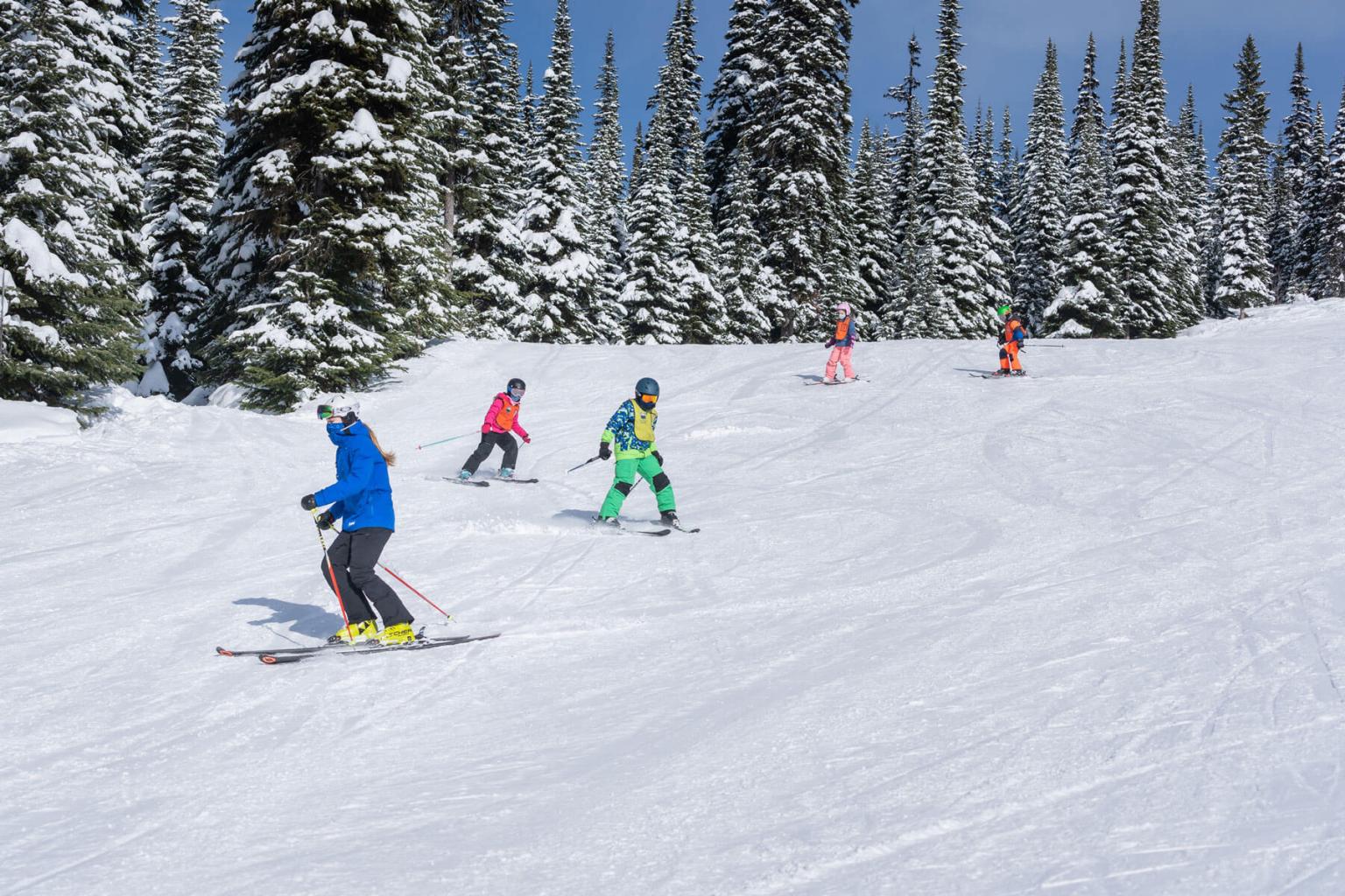 A ski instructor followed by four kids skiing down the run with trees in the background 