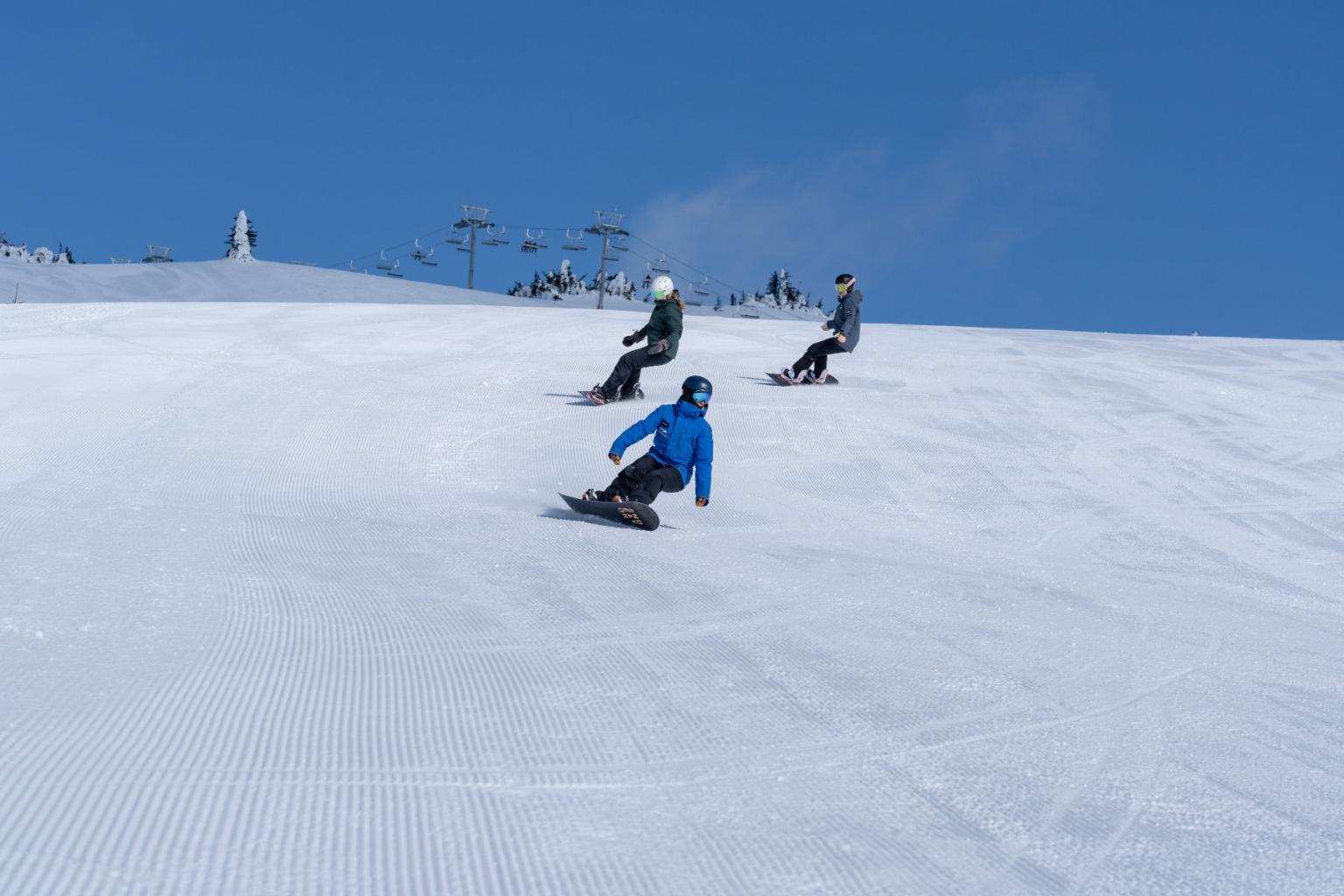 Three snowboarders riding down a run with the chairlift and blue skies behind them