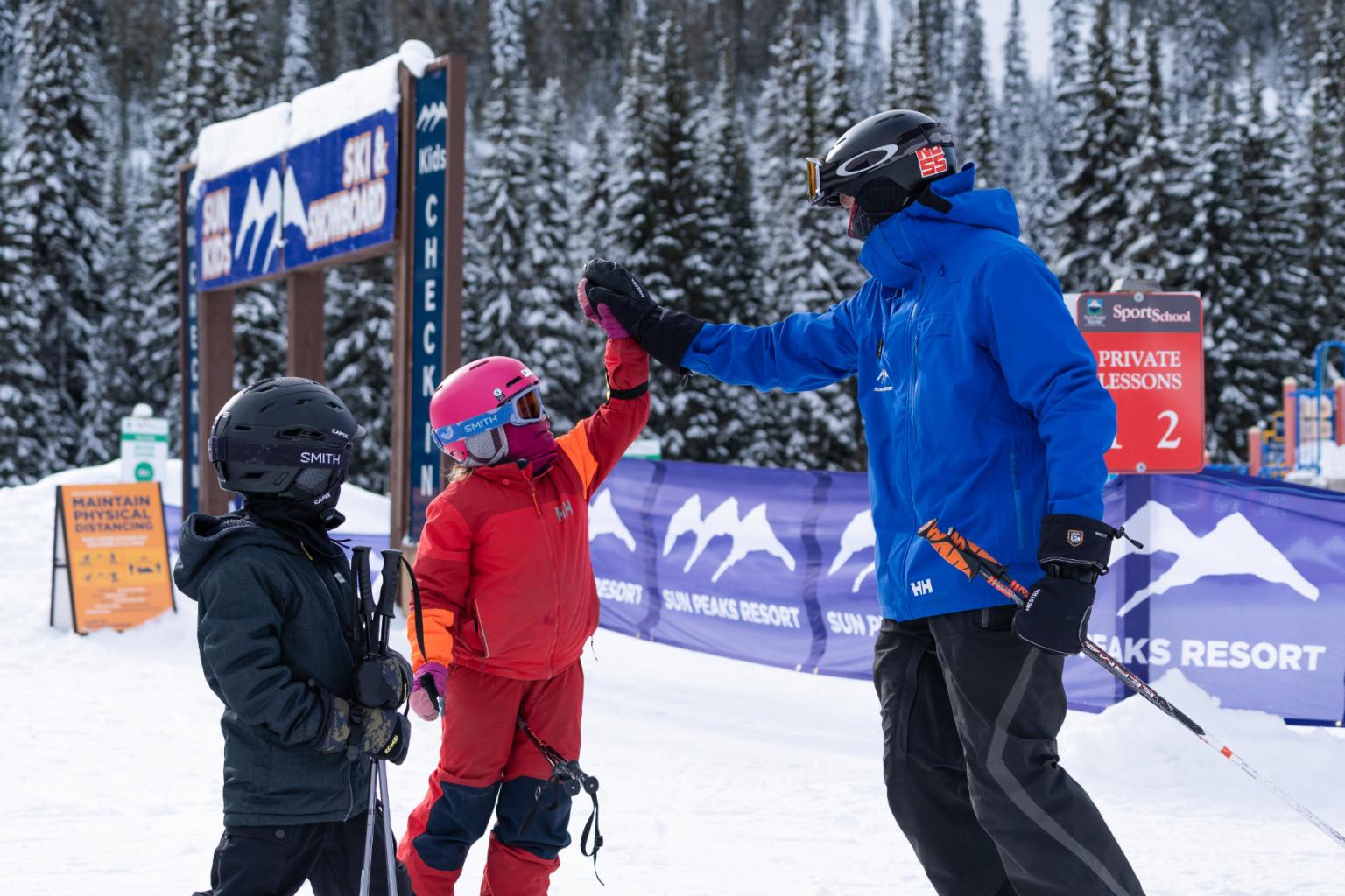 A ski instructor high fiving a younger skiers with trees and village signage in the background