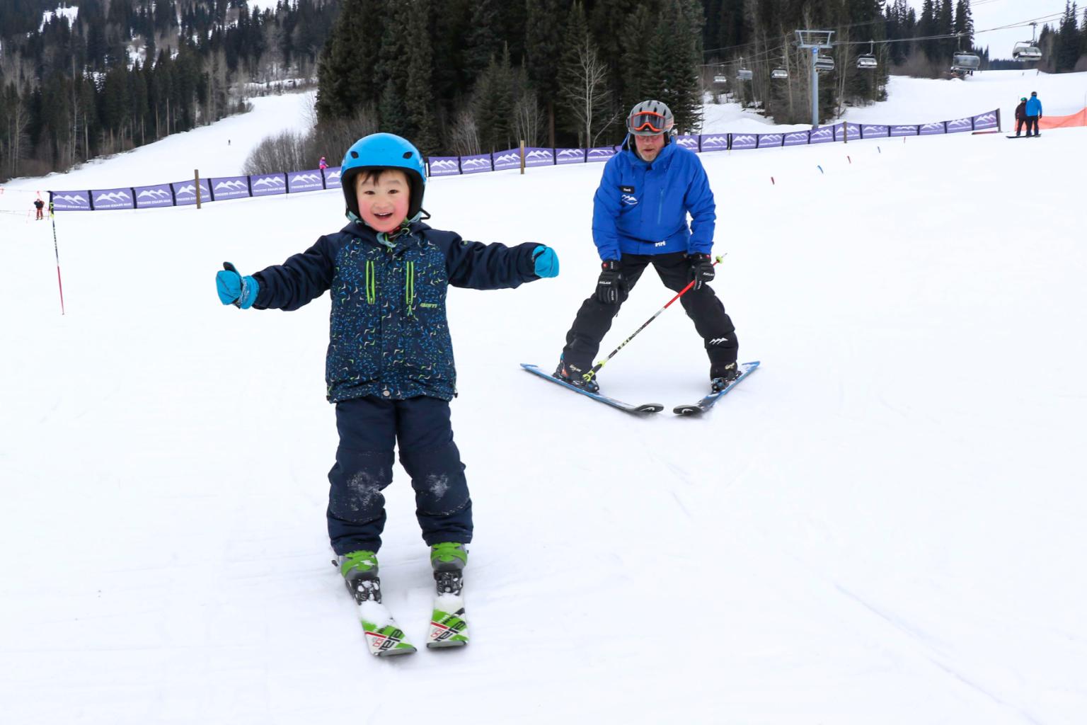 A child on skis skiing towards the camera with the instructor behind them with trees in the background