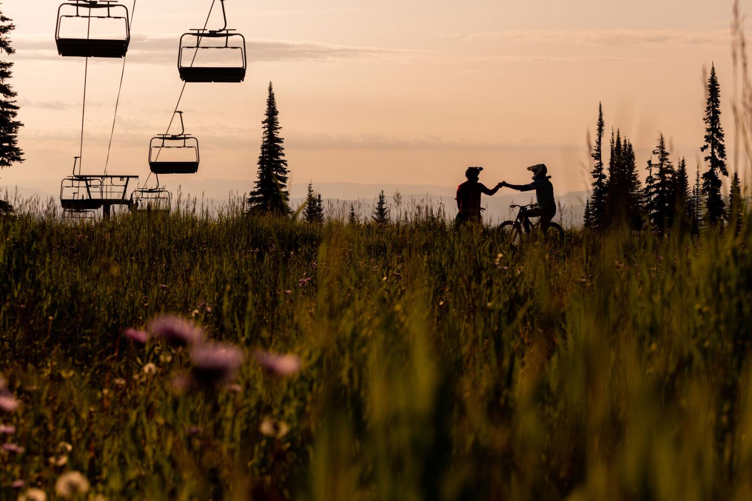 Two bikers in a field at sunset with a chairlift and mountain views
