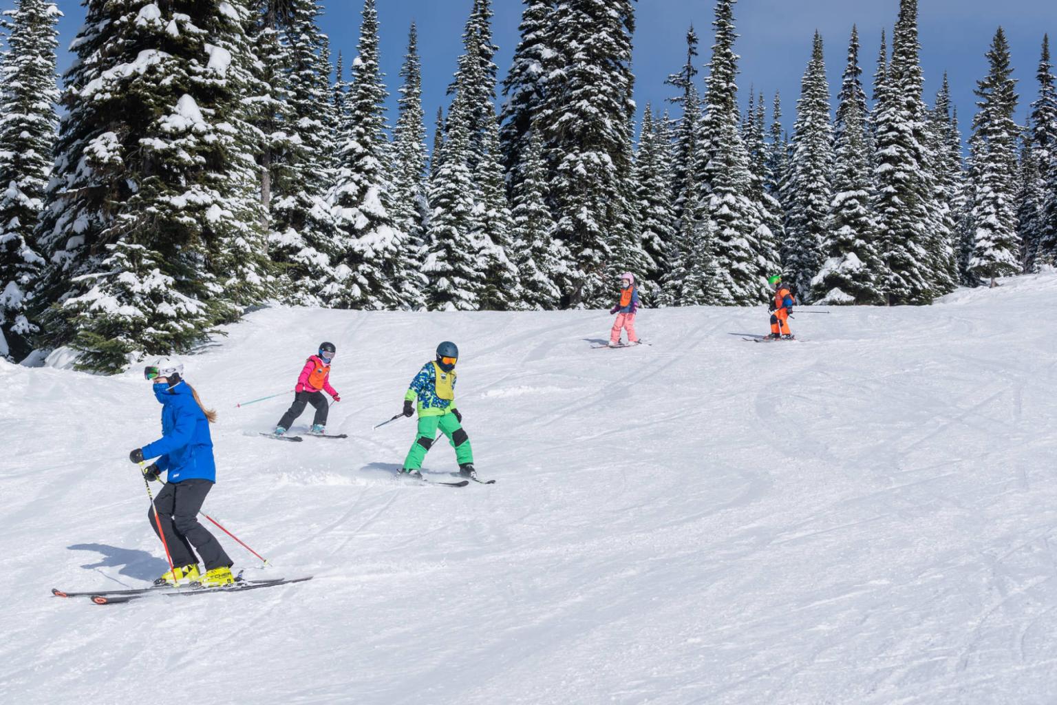 Ski instructor with kids following them down the run with trees behind them