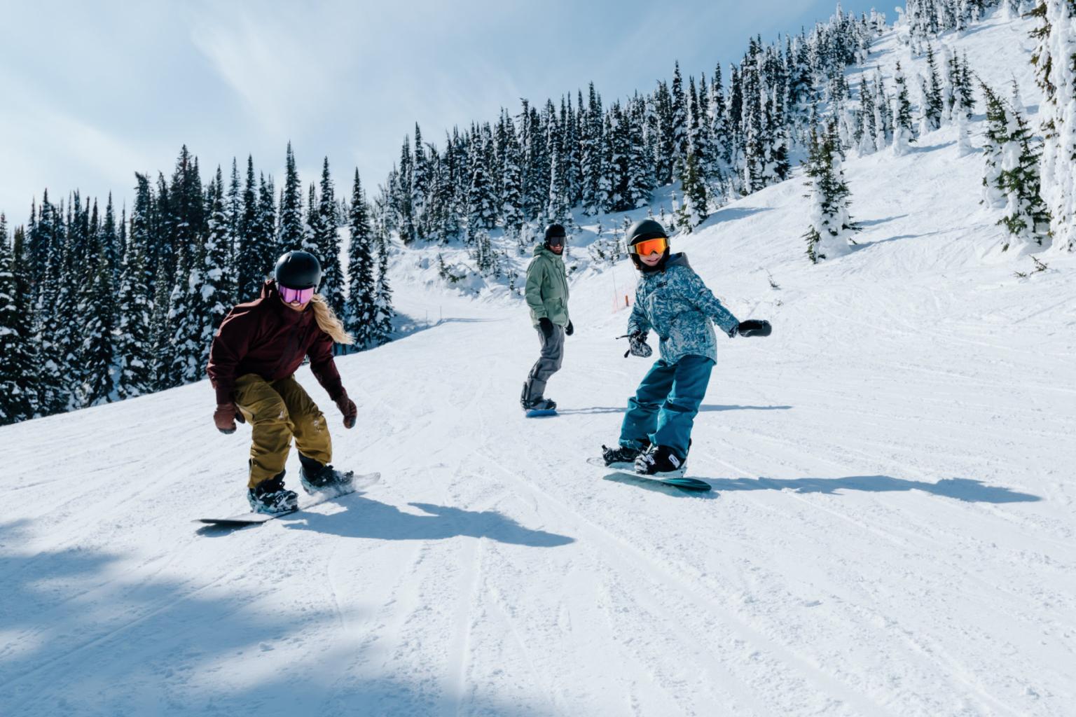 A group of 3 snowboarding down the mountain with trees and mountain views behind them