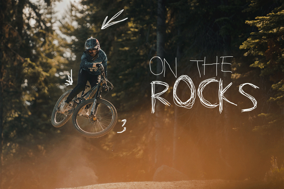 Woman whipping her bike over a large jump with a sun-flare over the photo and the title, "On the Rocks" in white