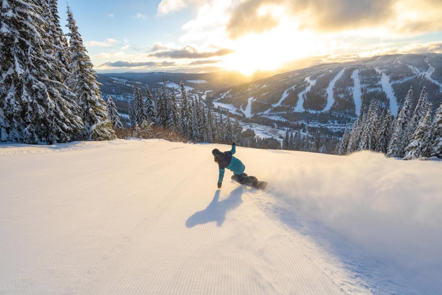 Snowboarder in blue jacket rides a powder covered groomer with the Village in the distance